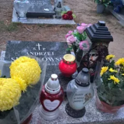 Uporządkowany nagrobek z szarego kamienia, ozdobiony żółtymi kwiatami, lampionami i wizerunkiem Jezusa. Tło stanowią inne groby i fragmenty drzew.