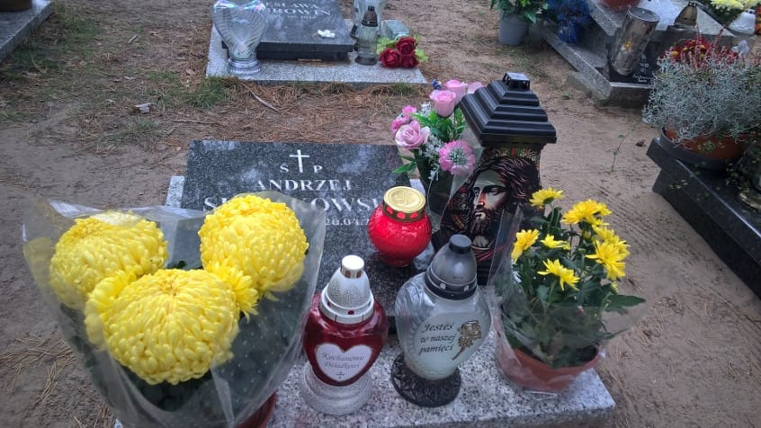 Uporządkowany nagrobek z szarego kamienia, ozdobiony żółtymi kwiatami, lampionami i wizerunkiem Jezusa. Tło stanowią inne groby i fragmenty drzew.