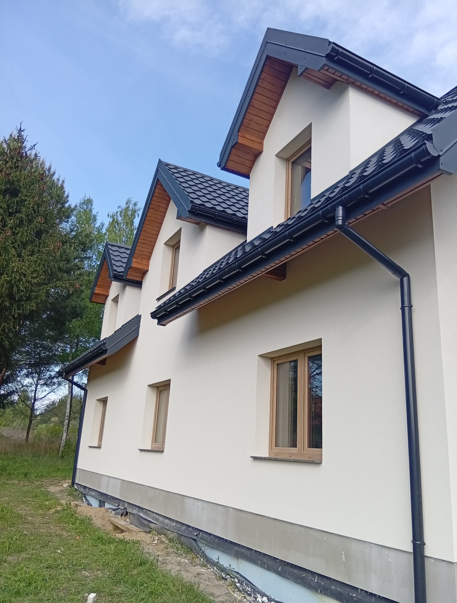 Nowoczesny dom z jasną elewacją, ciemną dachówką i drewnianymi elementami wykończeniowymi. Widoczne okna i rynny. Tło stanowi zieleń drzew i błękitne niebo.