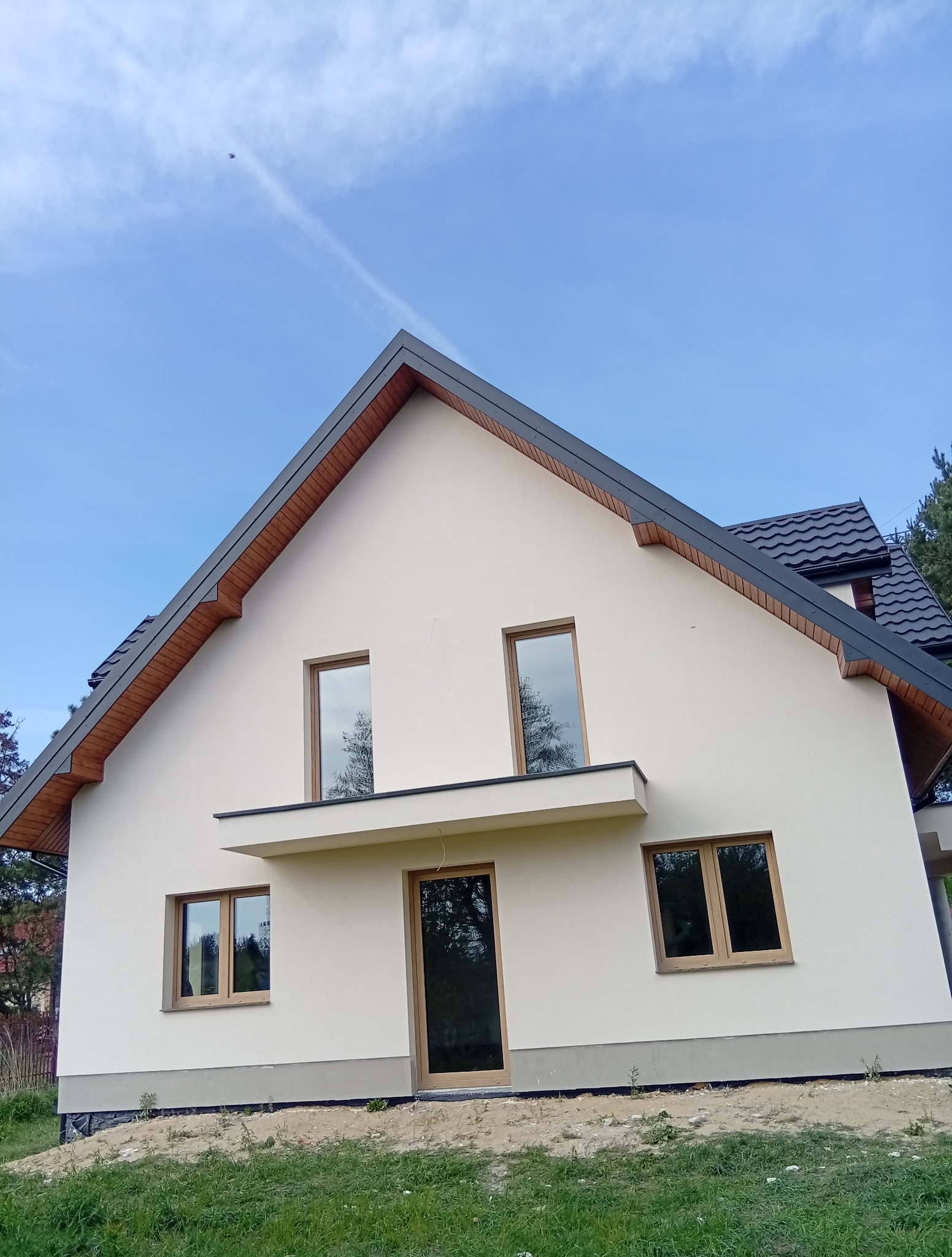 Jasna elewacja domu jednorodzinnego z drewnianymi oknami i ciemną dachówką na tle błękitnego nieba z widocznym śladem po samolocie.