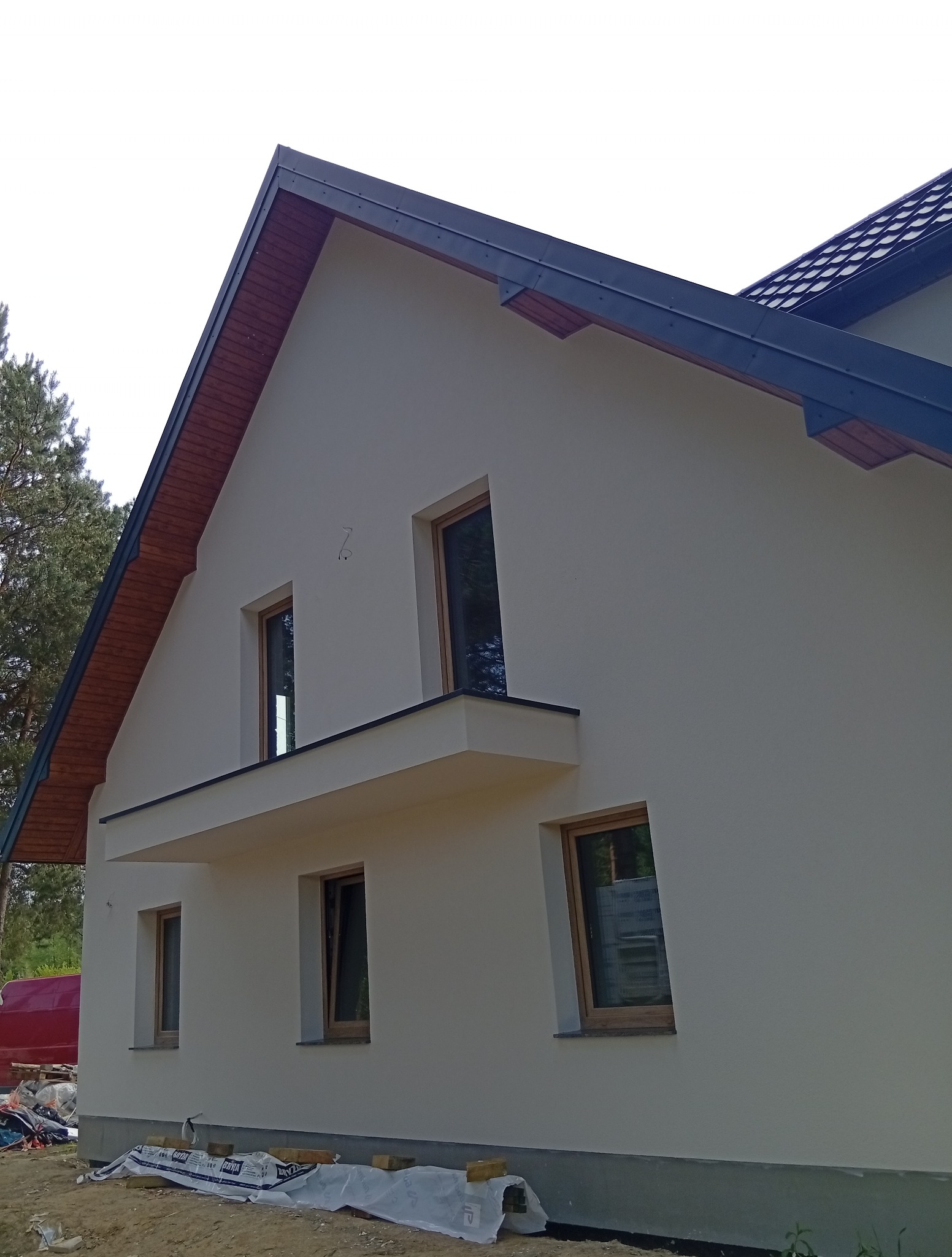 Jasna elewacja domu jednorodzinnego z drewnianymi oknami i grafitowym dachem. Widoczne materiały budowlane sugerują prace wykończeniowe.