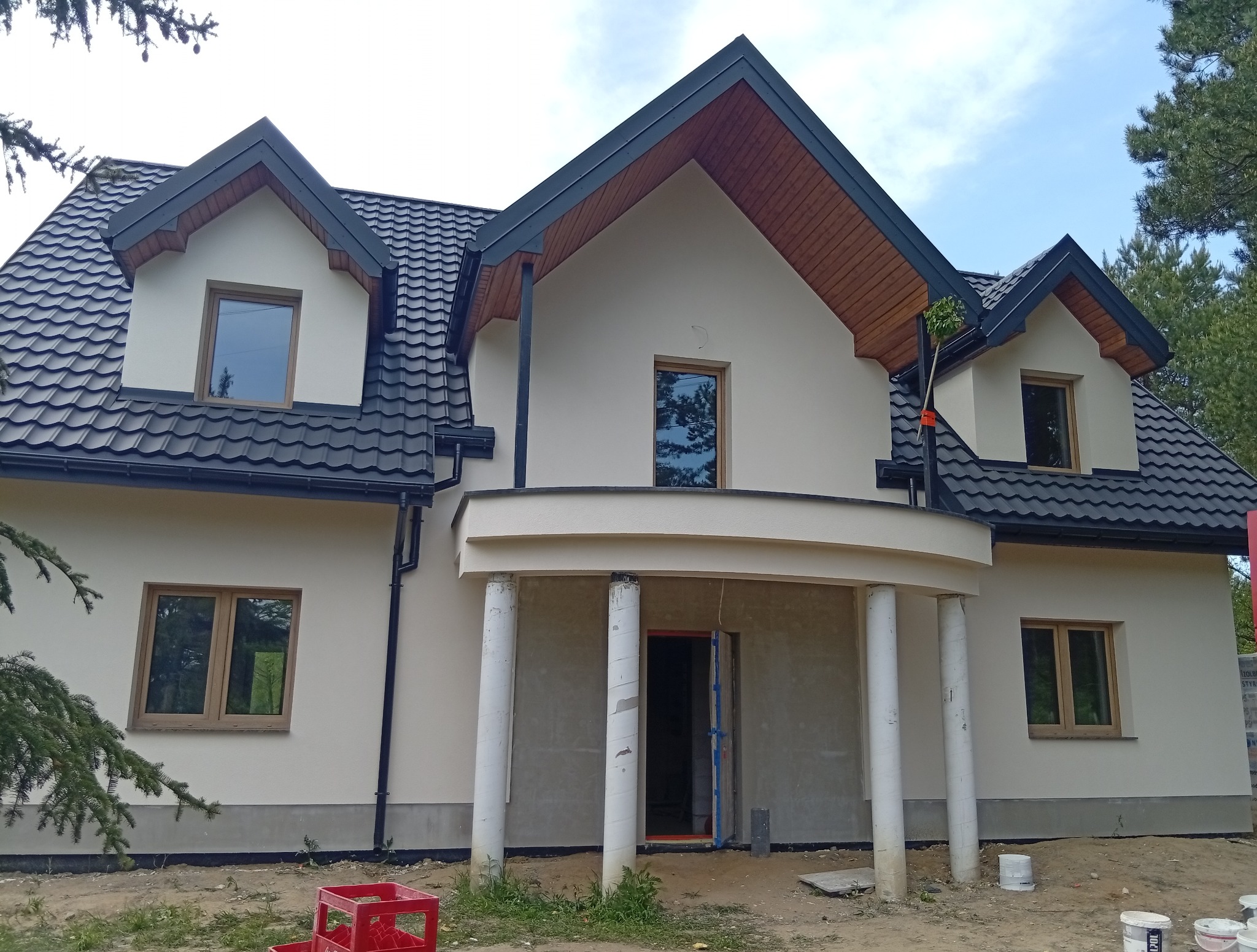Jasna elewacja domu z ciemnym dachem i drewnianymi elementami wykończeniowymi. Widoczne kolumny i niezakończone prace wykończeniowe.