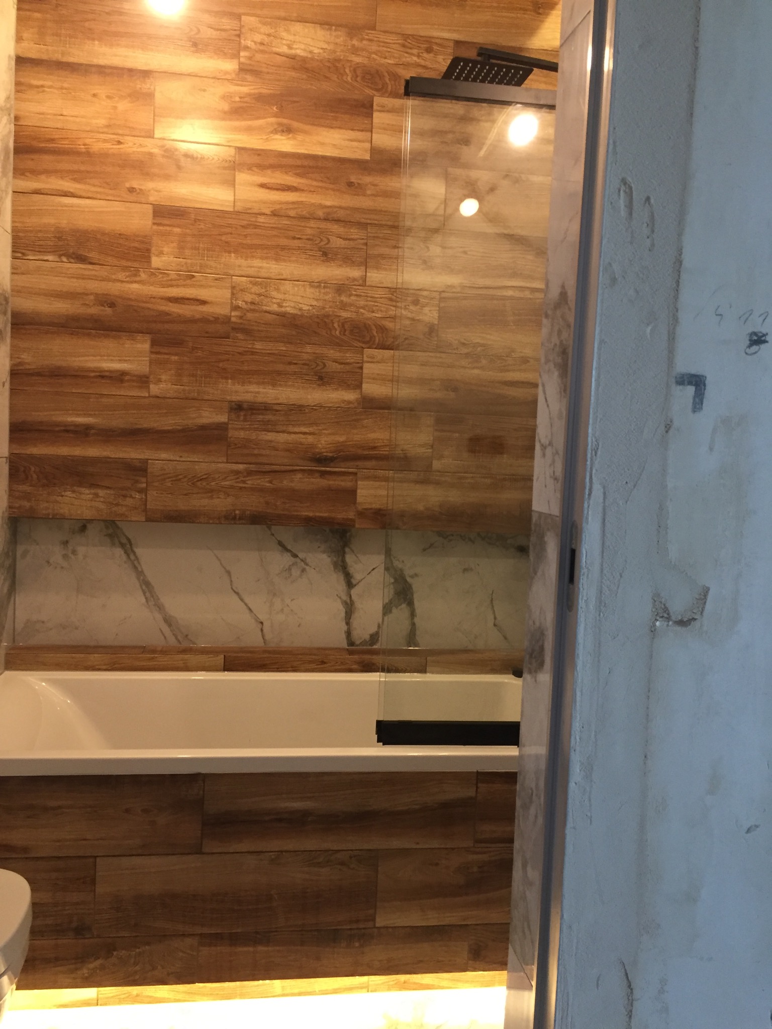 Wykończona łazienka z wanną obudowaną płytkami imitującymi drewno, nad nią fragment ściany z płytek marmurowych i szklana kabina prysznicowa.
