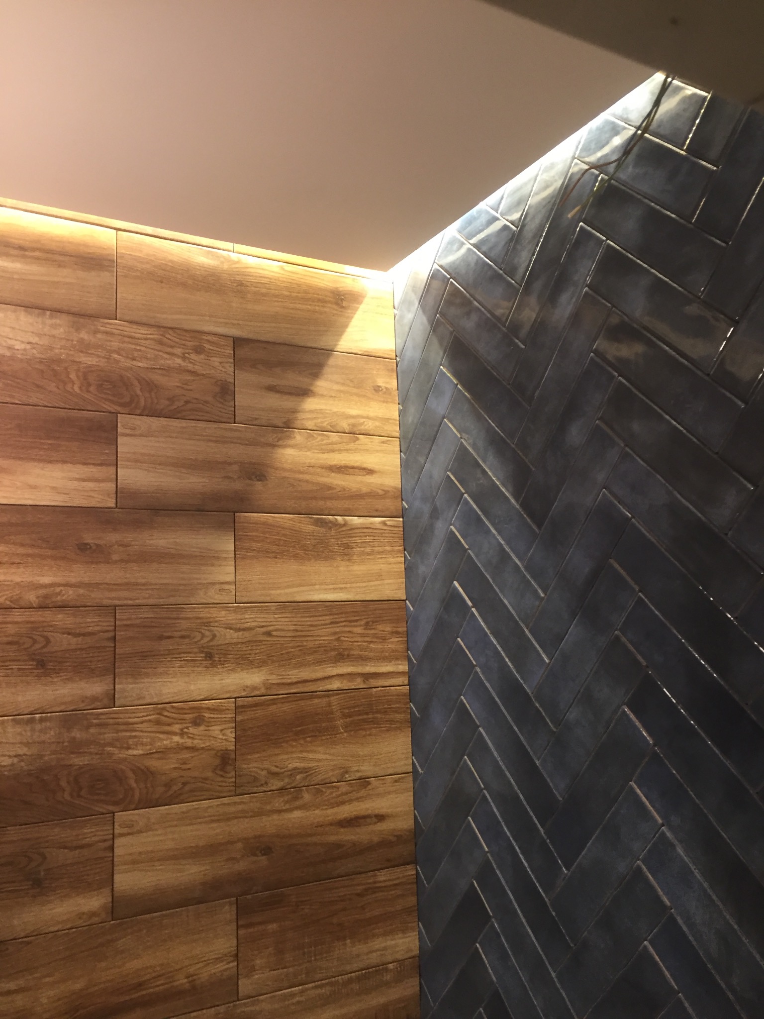 Ściana wykończona płytkami imitującymi drewno i ciemnymi płytkami ułożonymi w jodełkę, z subtelnym oświetleniem LED podkreślającym fakturę.