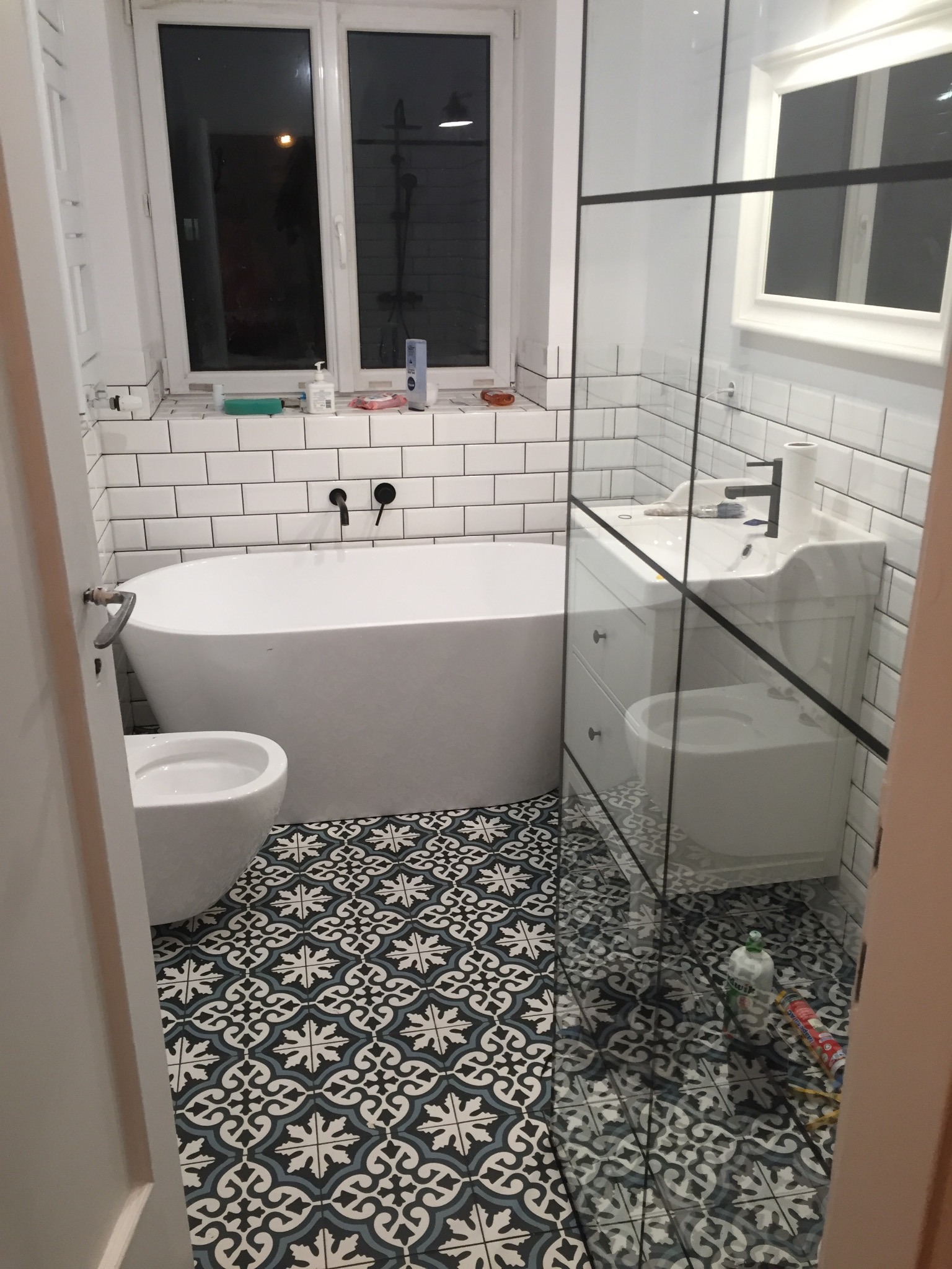 Nowoczesna łazienka z wolnostojącą wanną, białą armaturą i wzorzystą podłogą. Widoczne czarne baterie i szklana kabina prysznicowa. Świeże, jasne wnętrze.