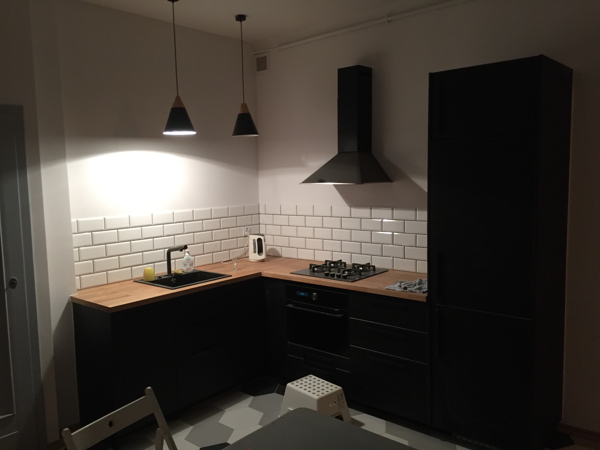 Nowoczesna kuchnia w czerni i drewnie: białe płytki, czarne szafki, drewniany blat, minimalistyczne lampy zwisające. Ukończona aranżacja wnętrza.