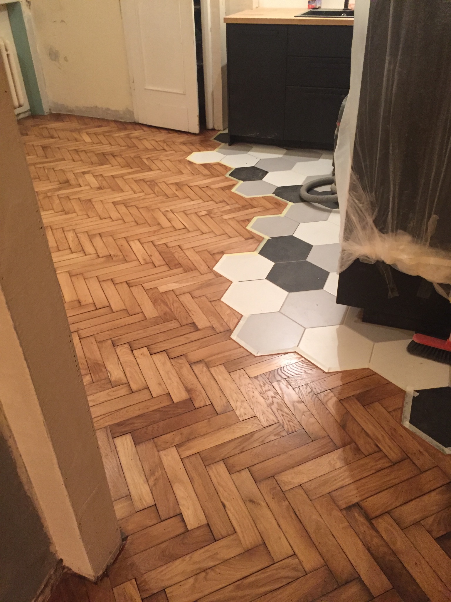 Parkiet ułożony w jodełkę przechodzi w heksagonalne płytki w odcieniach szarości. Widoczny fragment szafki kuchennej i odkurzacza. Częściowo ukończona podłoga.