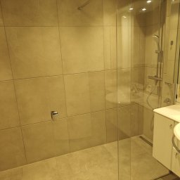 Renovation & Construction Zbigniew Bogdan - Nowoczesna kabina prysznicowa ze szklaną ścianą i deszczownicą w łazience wykończonej beżowymi płytkami. Widoczna umywalka z szafką.