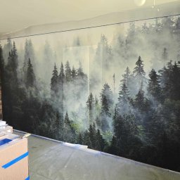 Renovation & Construction Zbigniew Bogdan - Tapeta z motywem mglistego lasu na ścianie, zabezpieczona folią malarską przy suficie. Widoczne narzędzia i materiały malarskie. Część ściany z cegły obok.