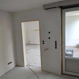 Renovation & Construction Zbigniew Bogdan - Wnętrze w trakcie prac wykończeniowych: świeżo wyszpachlowane ściany z widocznymi puszkami elektrycznymi, przygotowane do dalszych prac. Widoczne przejście do innego pomieszczenia.