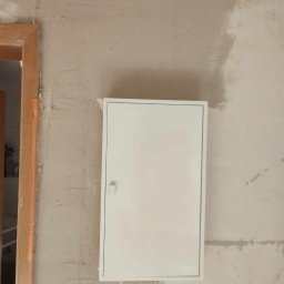 Renovation & Construction Zbigniew Bogdan - Biała skrzynka elektryczna na surowej ścianie po szpachlowaniu, widoczny fragment ościeżnicy drzwi. Ściana przygotowana do malowania.