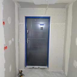 Renovation & Construction Zbigniew Bogdan - Wnętrze w trakcie prac wykończeniowych: szpachlowane ściany, osadzona metalowa futryna z drzwiami i narzędzia budowlane na podłodze.