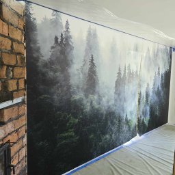 Renovation & Construction Zbigniew Bogdan - Ściana z fototapetą leśną, zabezpieczona folią i taśmą malarską, obok fragment ściany z cegły. Przygotowania do prac wykończeniowych.