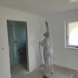 Renovation & Construction Zbigniew Bogdan - Pracownik w kombinezonie maluje ściany natryskowo w jasnym pomieszczeniu. Widoczne okno z folią ochronną, otwór drzwiowy i kartony. Praca wre, aby odświeżyć wnętrze.