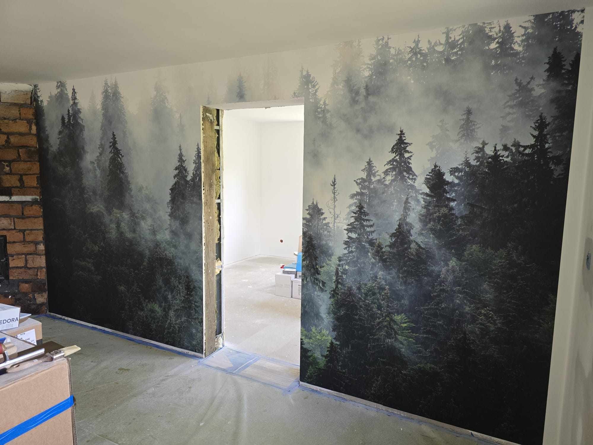 Fototapeta z motywem leśnym na ścianie, widok na sąsiednie pomieszczenie przez otwarte drzwi, w tle narzędzia malarskie i kartony.