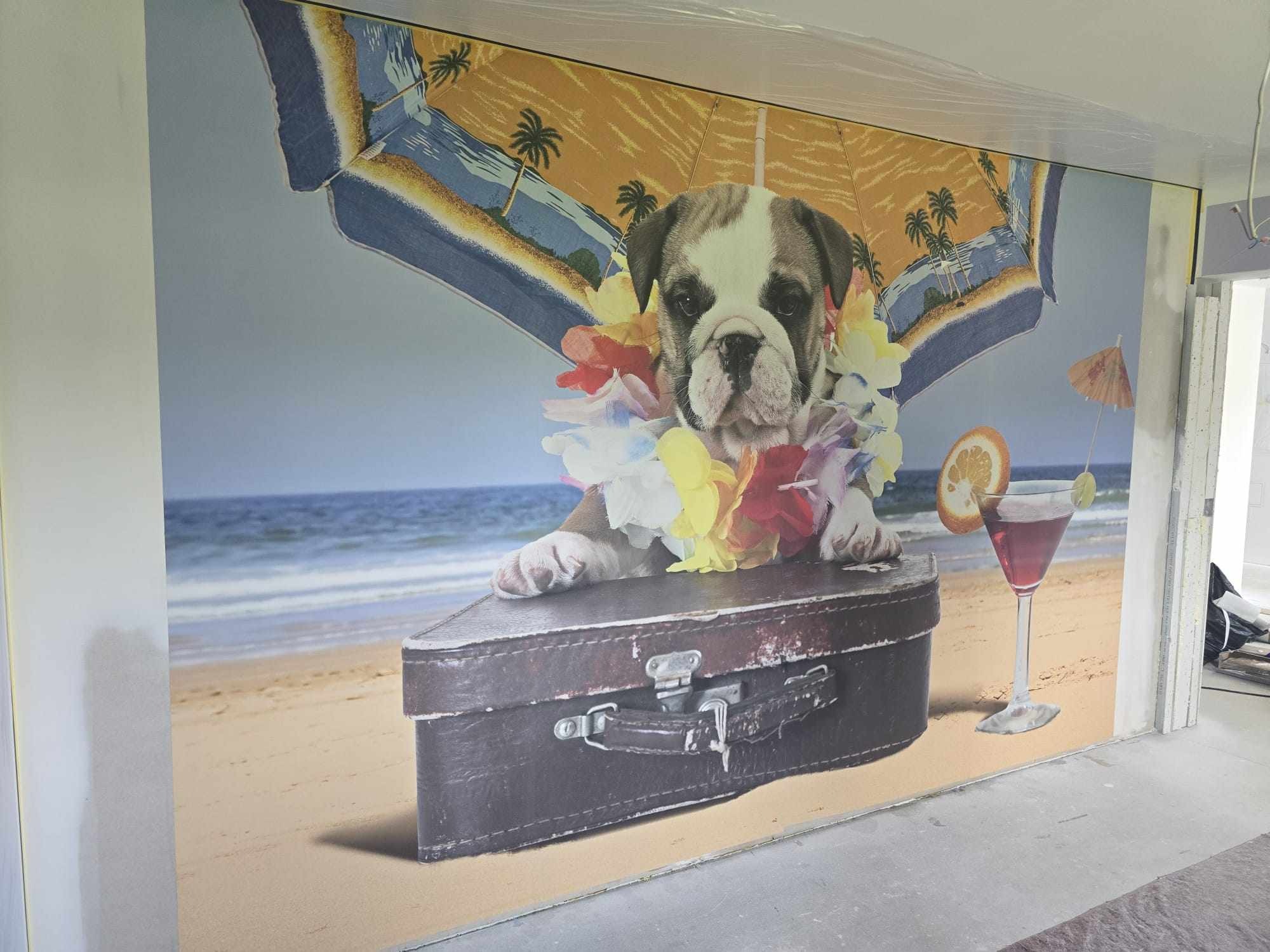 Fototapeta na ścianie z motywem psa w hawajskim wieńcu, drinka i walizki na tle plaży, z widocznymi elementami wykończeniowymi ściany.