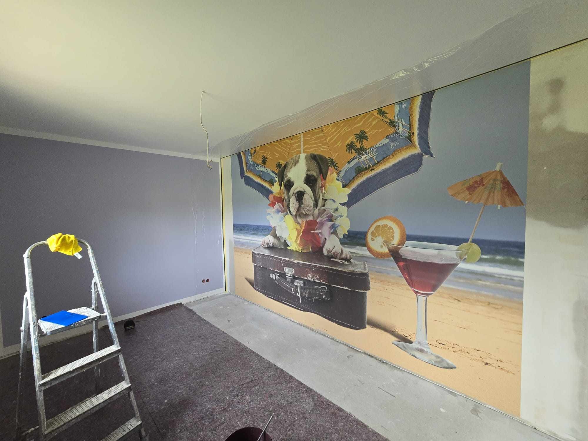 Pokój w trakcie malowania: drabina, zabezpieczona folią ściana z fototapetą z psem na plaży i koktajlem. Widoczne ślady prac malarskich.