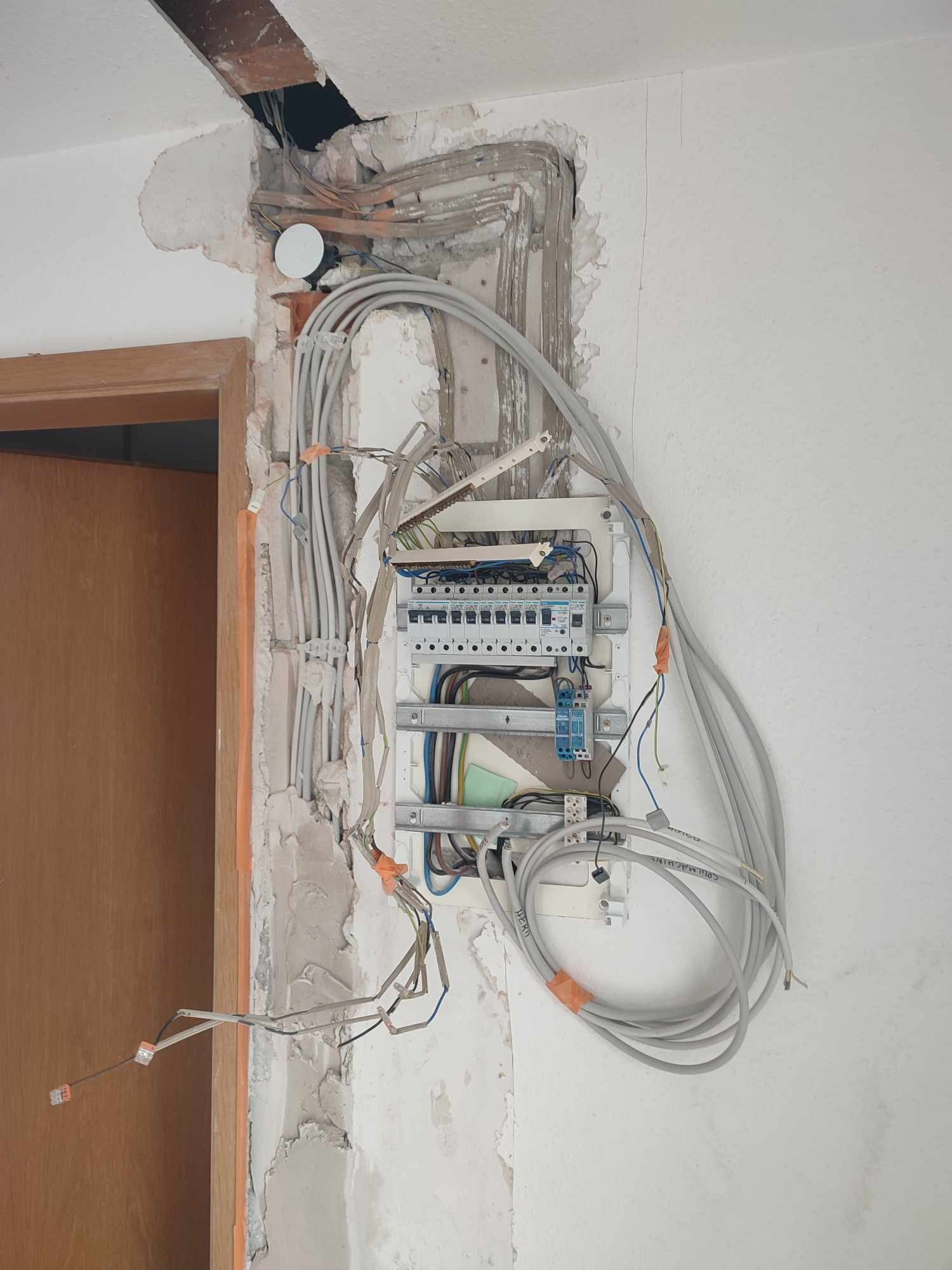 Rozdzielnia elektryczna w trakcie montażu, widoczne przewody, ściana w stanie surowym, obok ościeżnica drzwi. Instalacja elektryczna w mieszkaniu.