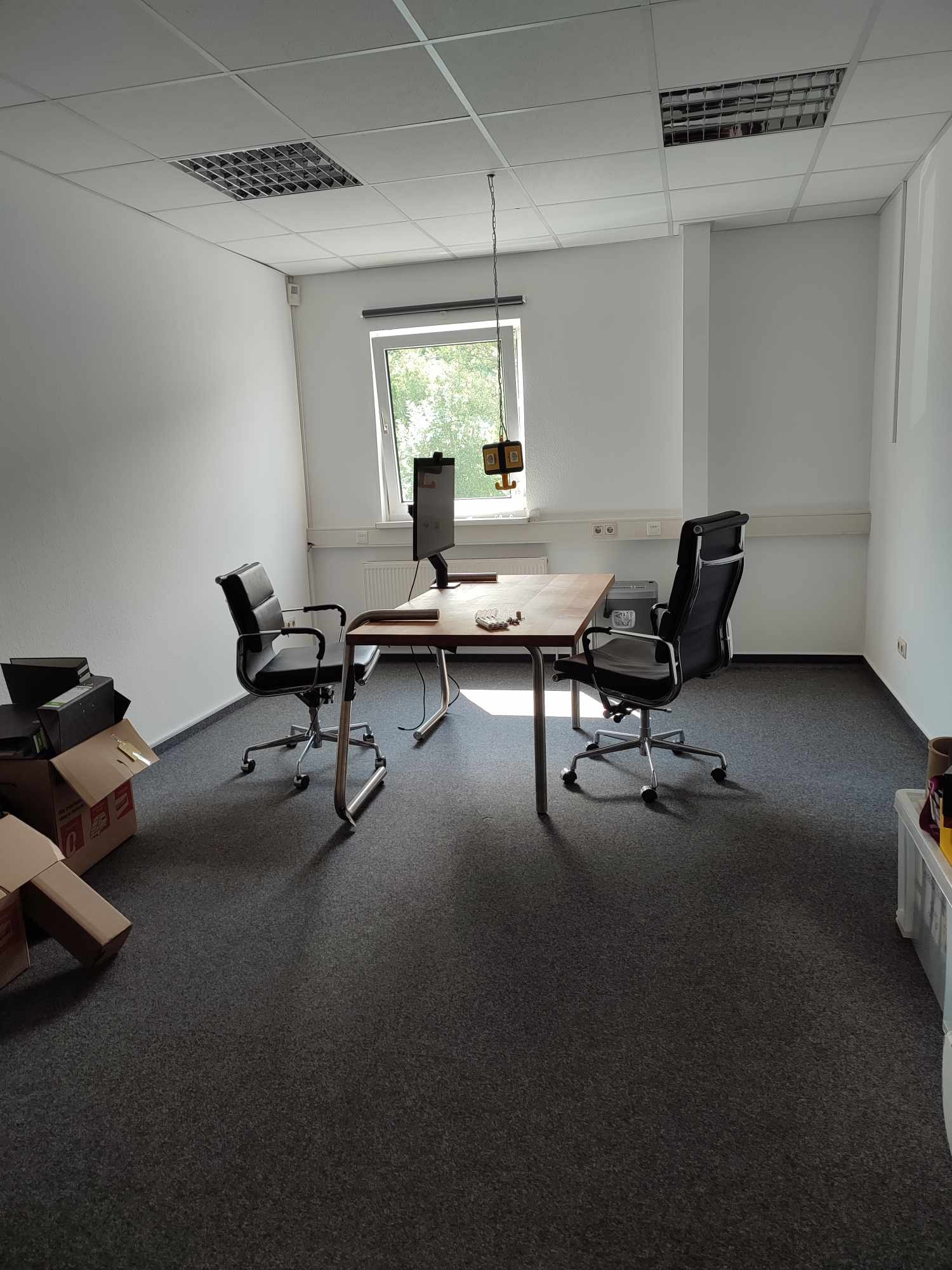 Wnętrze biura po remoncie: biurko z komputerem, krzesła obrotowe, szara wykładzina dywanowa, białe ściany, kartony w rogu. Widok na odnowione, jasne pomieszczenie.