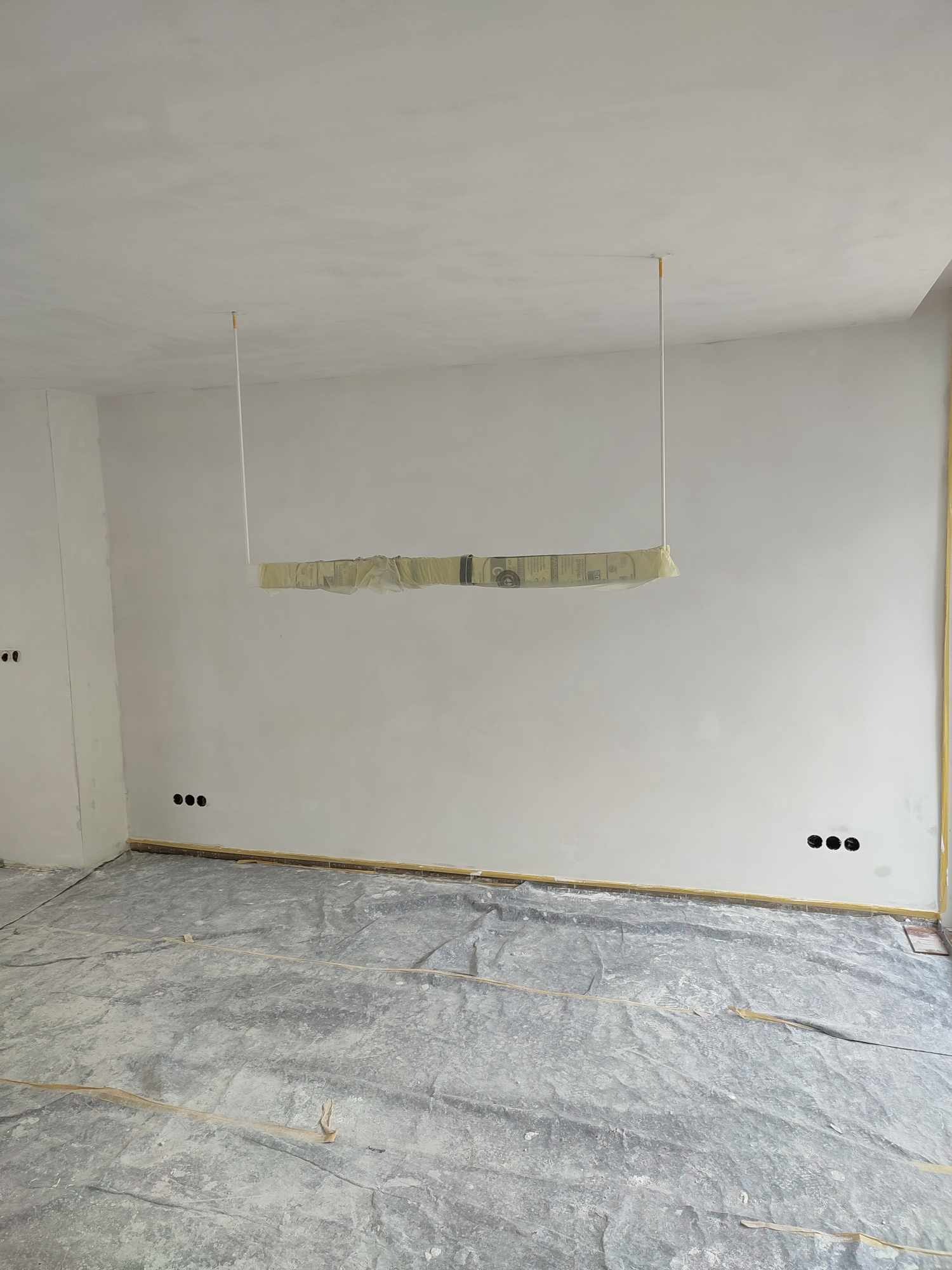 Wnętrze w trakcie malowania: świeżo pomalowane ściany na biało, zabezpieczona folią podłoga, widoczne puszki elektryczne i osłonięta lampa sufitowa.