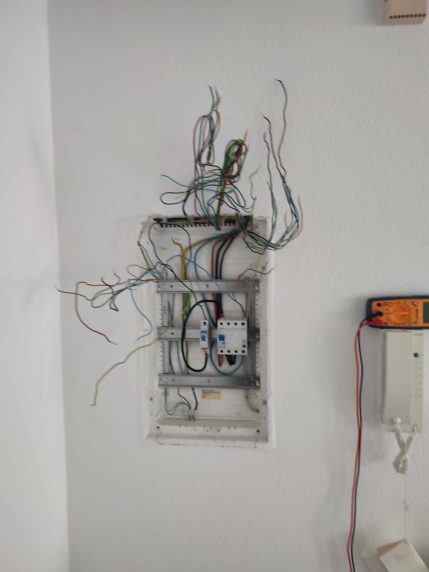 Otwarta skrzynka elektryczna na białej ścianie z wystającymi przewodami i podłączonym miernikiem. Widoczny fragment domofonu.