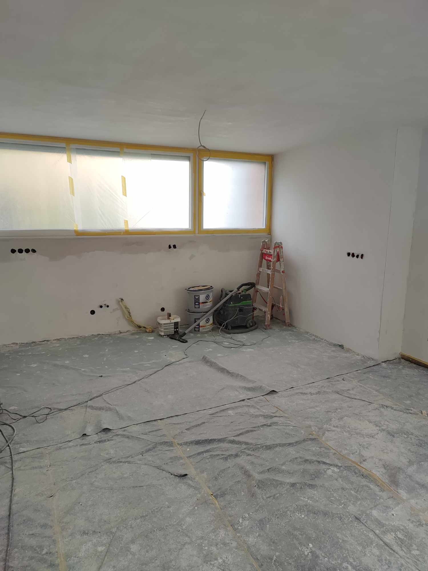 Wnętrze w trakcie przygotowań do malowania: okna zabezpieczone folią, podłoga przykryta, widoczne puszki z farbą, drabina i odkurzacz przemysłowy.