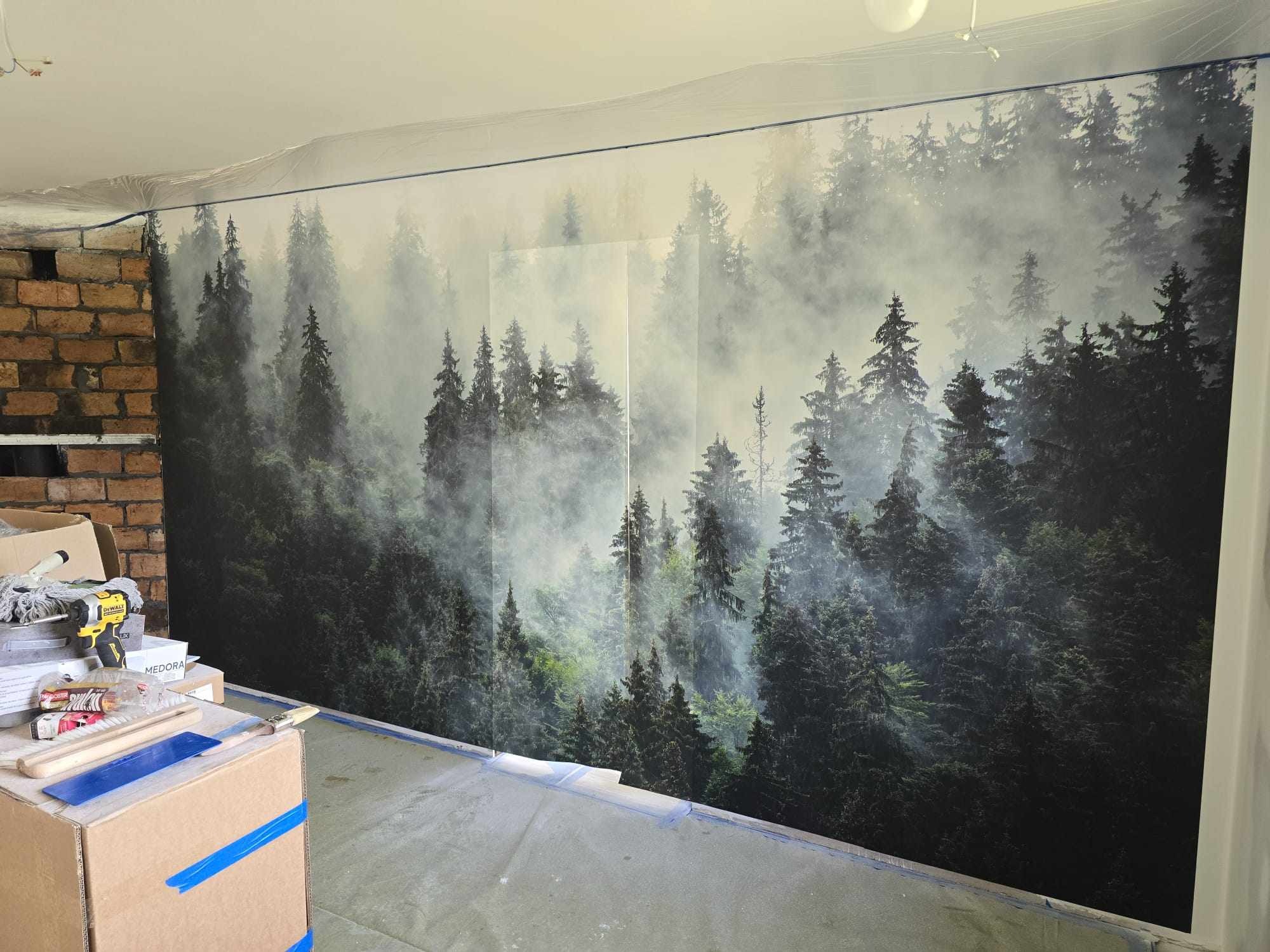 Tapeta z motywem mglistego lasu na ścianie, zabezpieczona folią malarską przy suficie. Widoczne narzędzia i materiały malarskie. Część ściany z cegły obok.