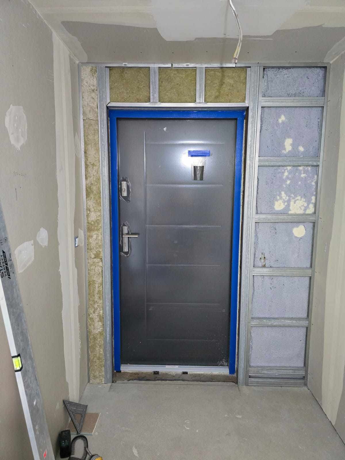 Montaż ościeżnicy metalowej z izolacją wokół szarych drzwi wejściowych. Widoczne profile, wełna mineralna i częściowo wykończone ściany gipsowe w trakcie prac.