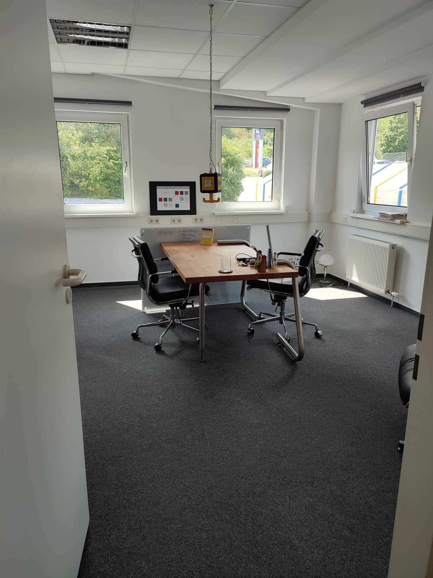 Wnętrze biura po remoncie w Brzostku: szary dywan, drewniany stół, czarne krzesła, białe ściany i okna z widokiem na zieleń. Minimalistyczny design, gotowe do użytku.