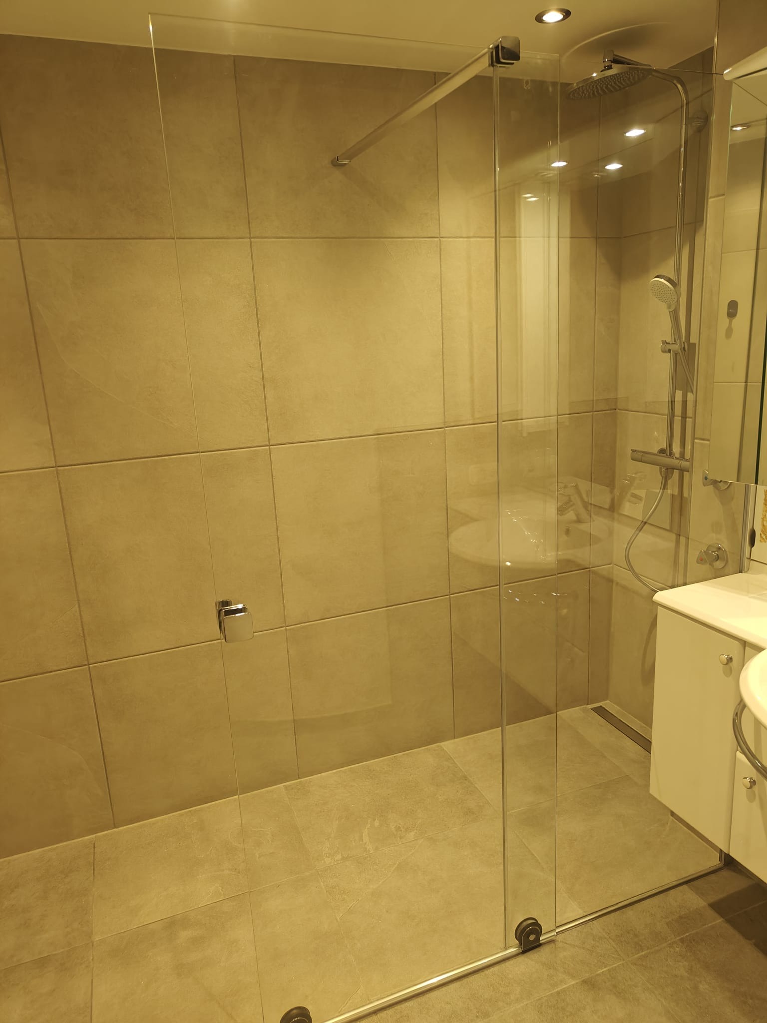 Nowoczesna łazienka z kabiną prysznicową typu walk-in, wykończona płytkami w odcieniach beżu i szarości, z widoczną armaturą i umywalką z szafką.