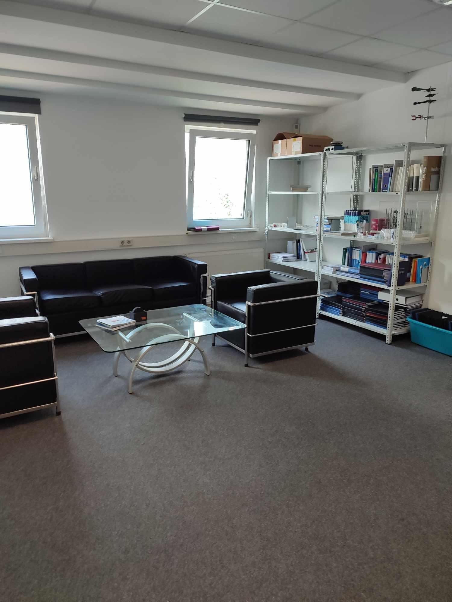 Jasne biuro z czarną skórzaną sofą, krzesłami i szklanym stolikiem kawowym. Metalowy regał z książkami i dokumentami przy ścianie. Dwa okna z roletami.