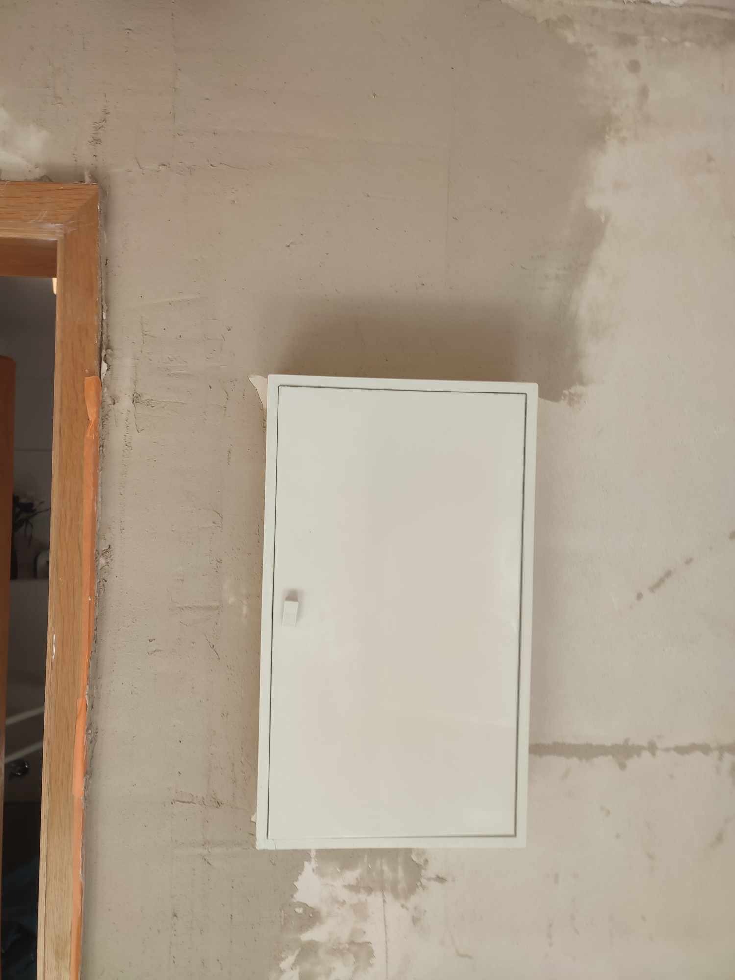 Biała skrzynka elektryczna na surowej ścianie po szpachlowaniu, widoczny fragment ościeżnicy drzwi. Ściana przygotowana do malowania.