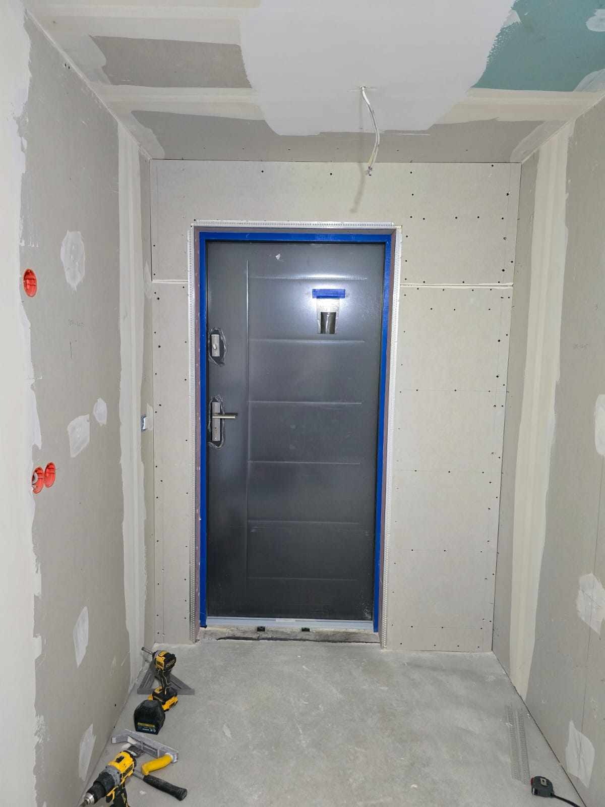 Wnętrze w trakcie prac wykończeniowych: szpachlowane ściany, osadzona metalowa futryna z drzwiami i narzędzia budowlane na podłodze.