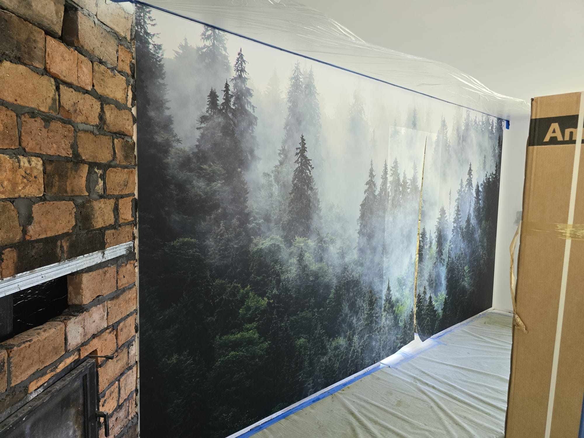 Ściana z fototapetą leśną, zabezpieczona folią i taśmą malarską, obok fragment ściany z cegły. Przygotowania do prac wykończeniowych.