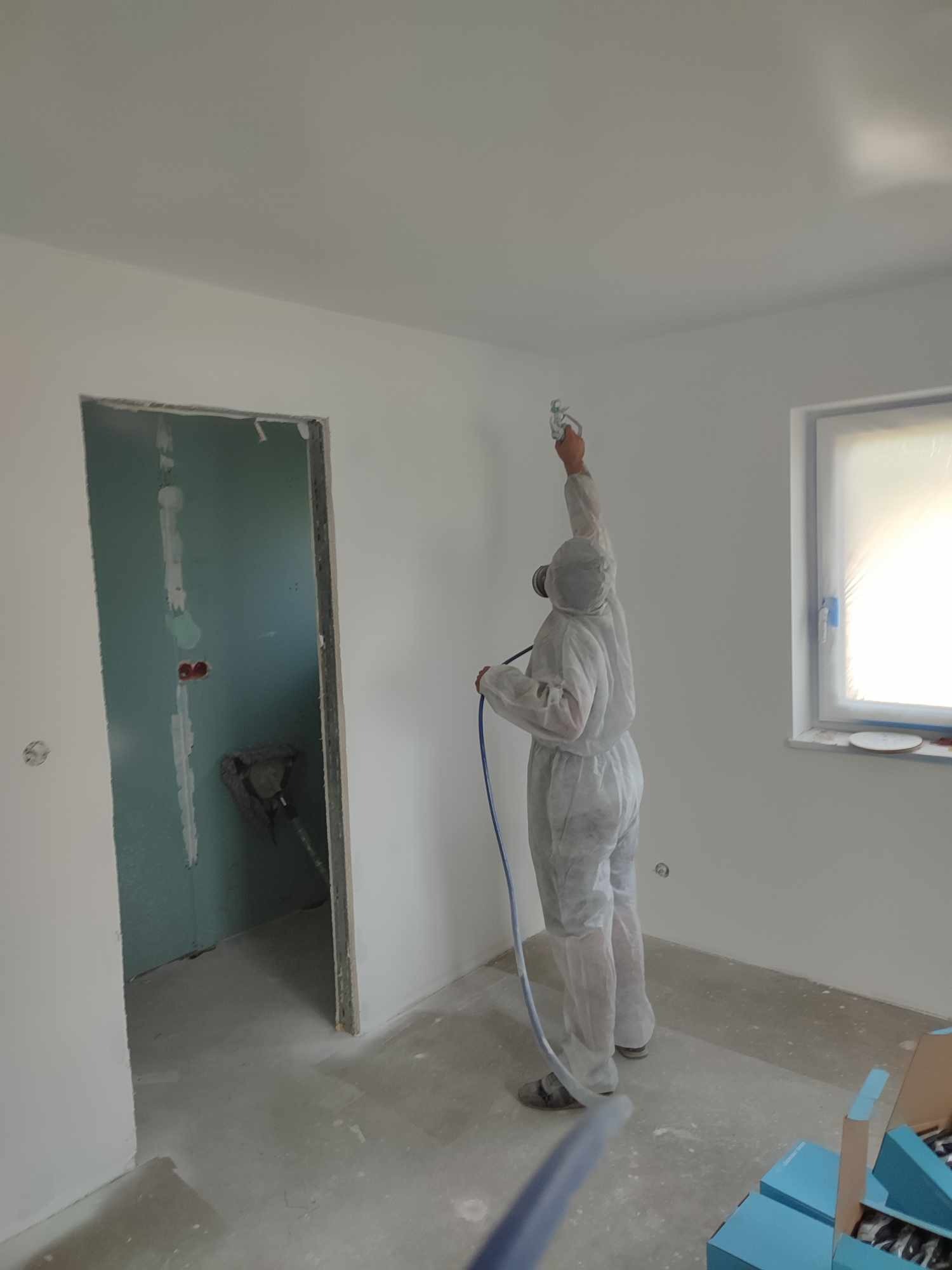 Pracownik w kombinezonie maluje ściany natryskowo w jasnym pomieszczeniu. Widoczne okno z folią ochronną, otwór drzwiowy i kartony. Praca wre, aby odświeżyć wnętrze.