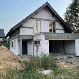 Inwestycje pod kontrolą - Surowy stan domu z garażem w Parsowie, z widocznymi oknami i dachem z dachówki, otoczony trawą i piaskiem. Budynek w trakcie budowy.