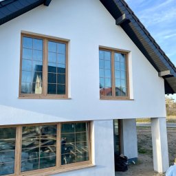 Inwestycje pod kontrolą - Nowoczesny dom z białymi ścianami i drewnianą stolarką okienną, czarnym dachem. W oknach odbija się otoczenie i wnętrze budynku. Widoczne kolumny podtrzymujące.