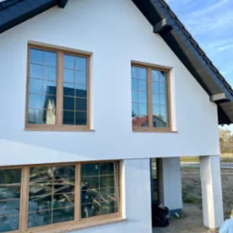 Nowoczesny dom z białymi ścianami i drewnianą stolarką okienną, czarnym dachem. W oknach odbija się otoczenie i wnętrze budynku. Widoczne kolumny podtrzymujące.