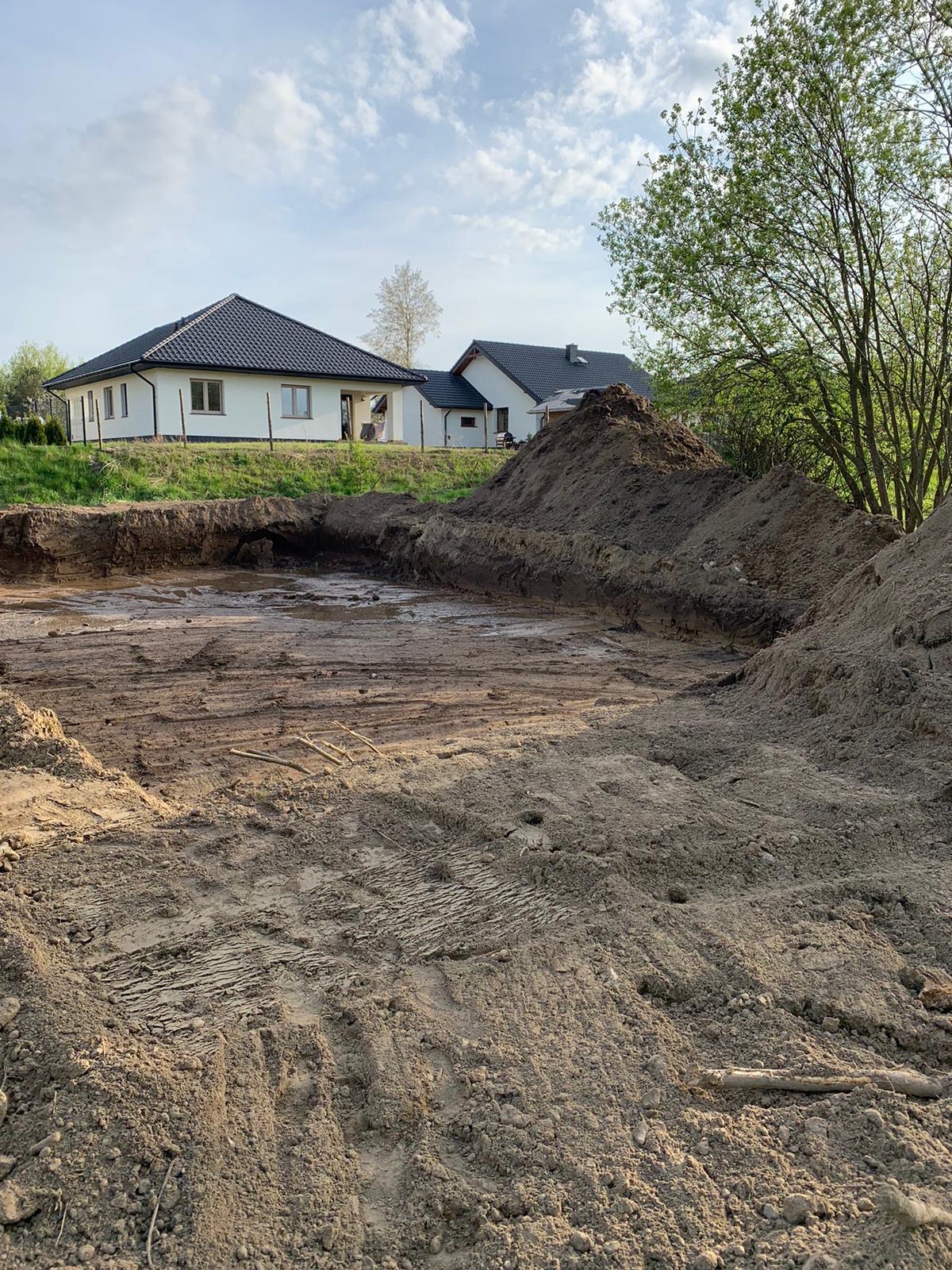 Wykonane wykopy pod fundamenty domu jednorodzinnego. W tle widoczne dwa budynki w stanie surowym zamkniętym z czarną dachówką i białymi elewacjami.