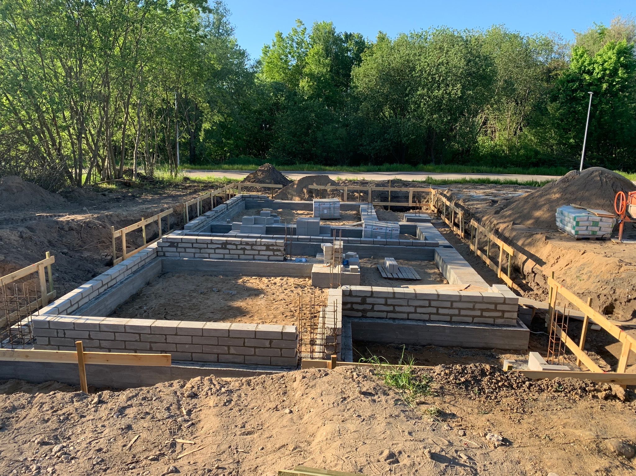 Fundamenty domu w trakcie budowy, widok z góry. Częściowo wymurowane ściany z bloczków, zbrojenia. W tle zielone drzewa i niebieskie niebo.