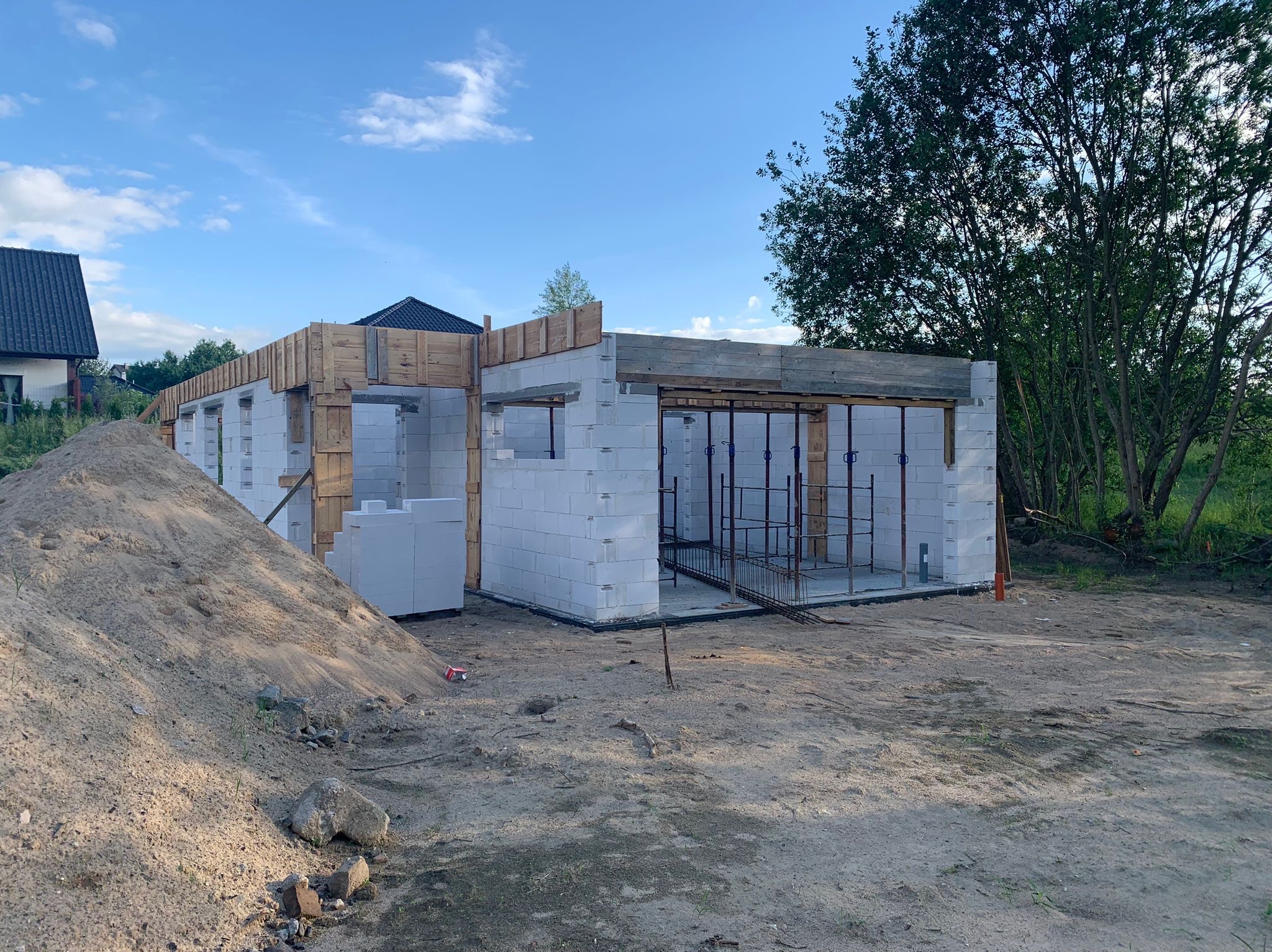 Budowa domu z betonu komórkowego w trakcie realizacji, widoczne zbrojenia i drewniane szalunki, hałda piasku na pierwszym planie, dom w tle, błękitne niebo.