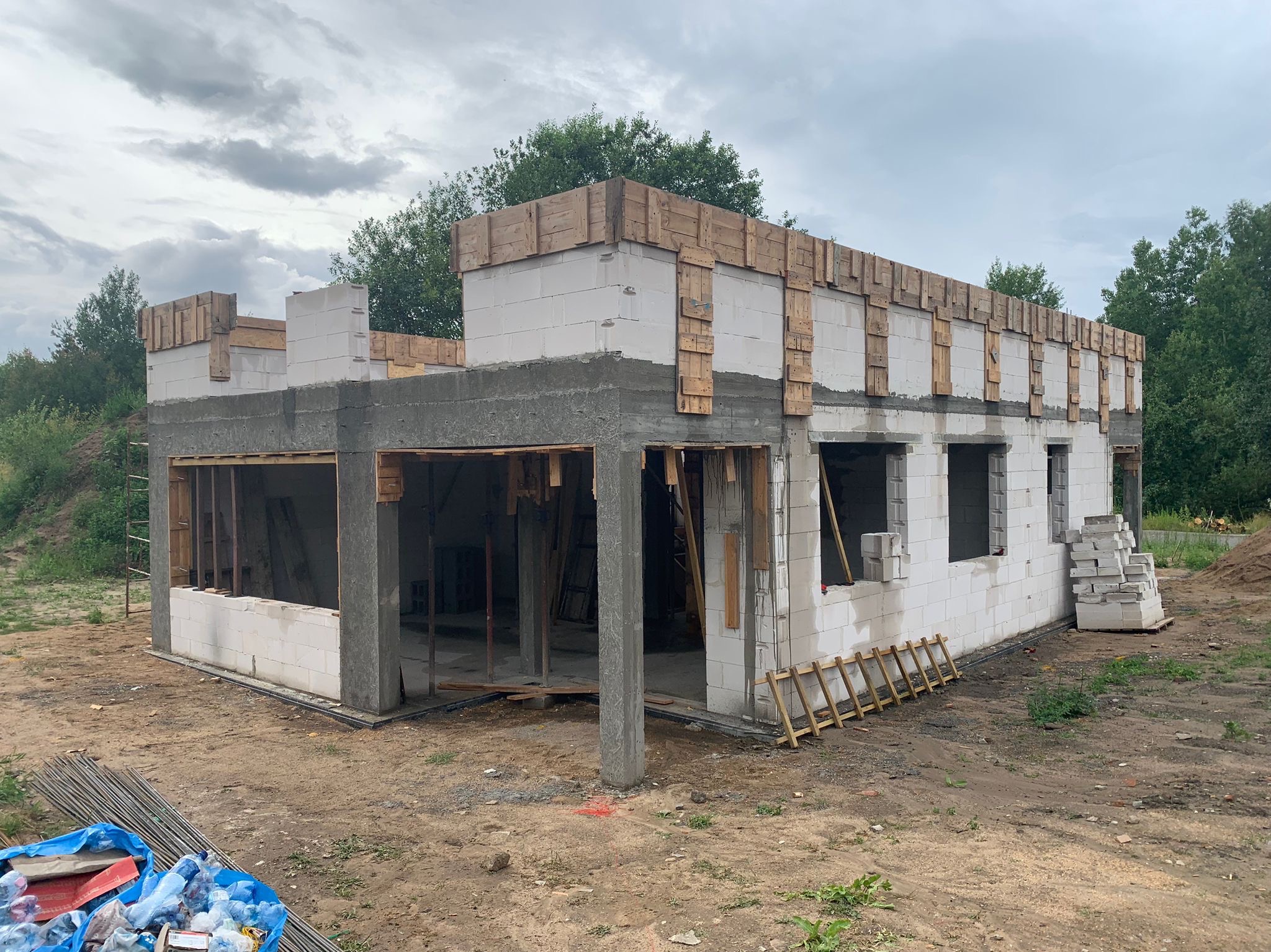 Budowa domu jednorodzinnego w stanie surowym otwartym: ściany z betonu komórkowego, drewniane szalunki, widoczne zbrojenia i drabina oparta o ścianę.