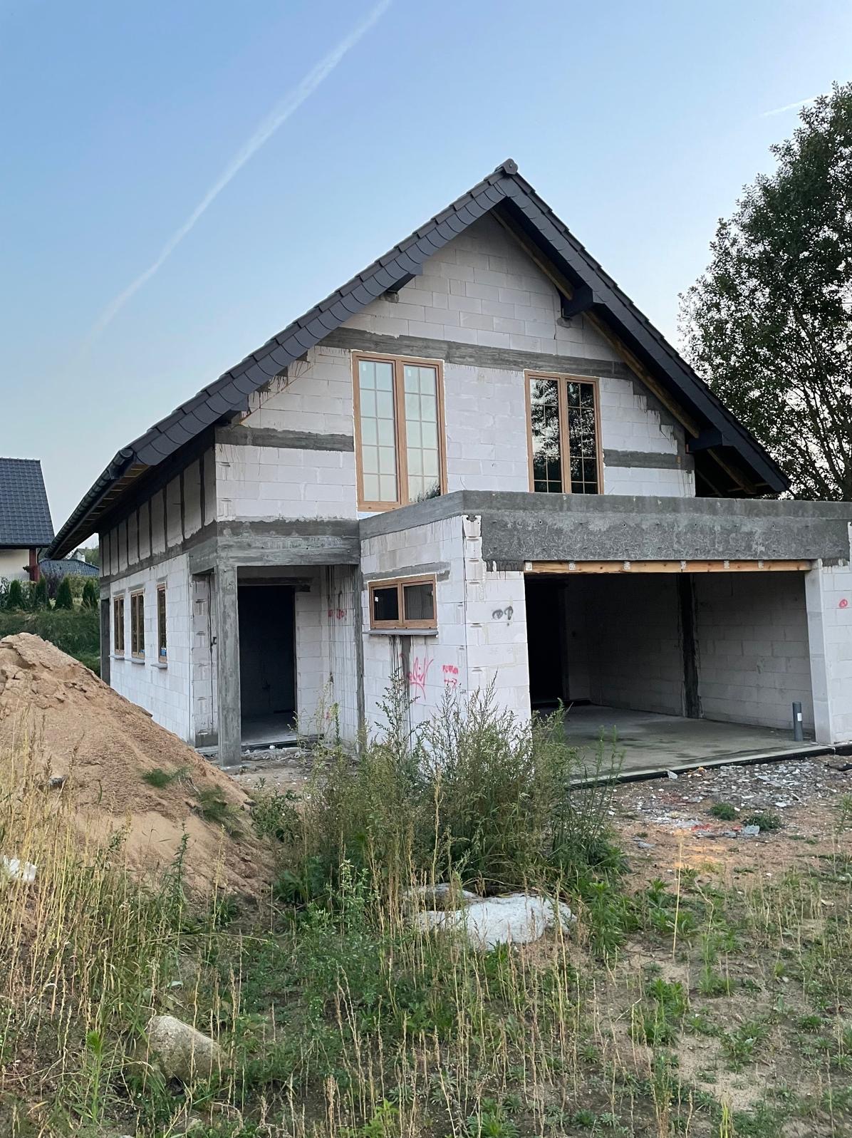 Surowy stan domu z garażem w Parsowie, z widocznymi oknami i dachem z dachówki, otoczony trawą i piaskiem. Budynek w trakcie budowy.
