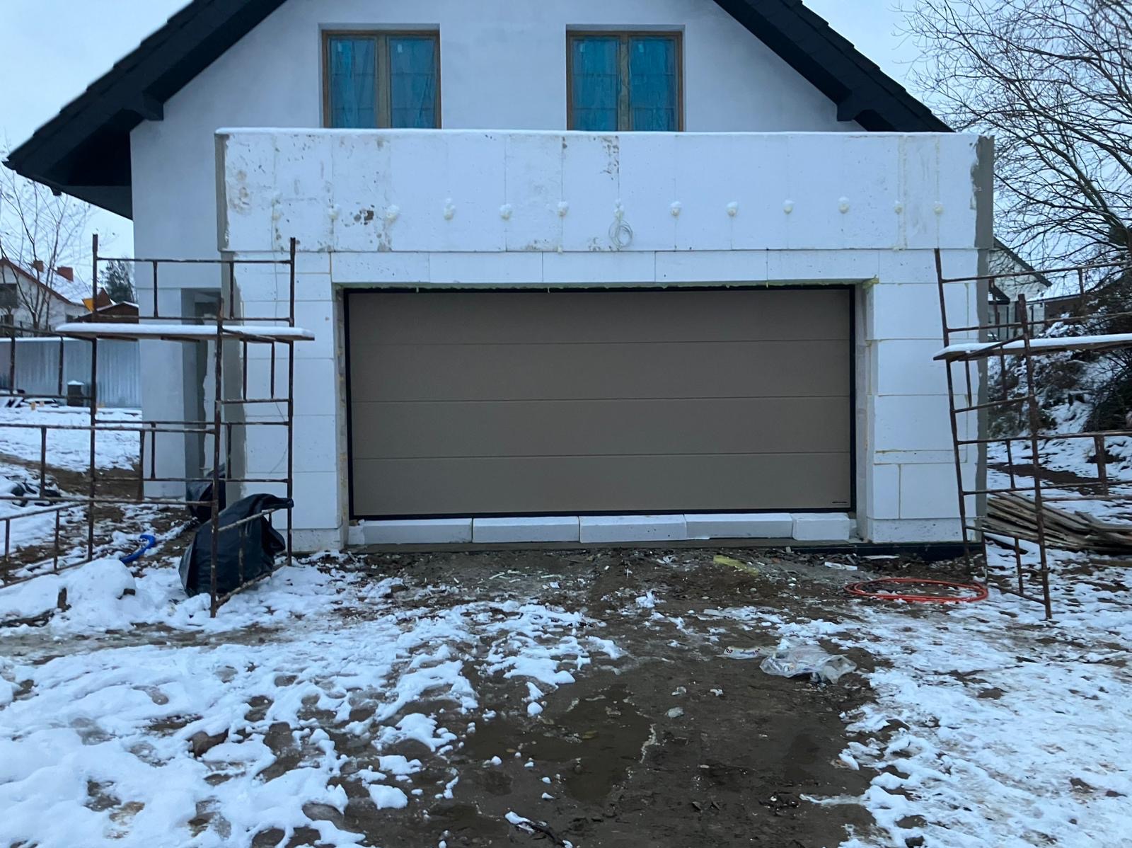 Montaż segmentowej bramy garażowej w nowym budynku z białego bloczka, widoczne rusztowania i resztki śniegu na ziemi.