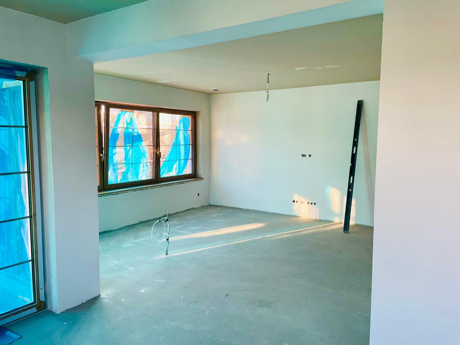 Surowe wnętrze w trakcie prac wykończeniowych: okna zabezpieczone folią, ściany po gładzi, widoczny poziomica i wystające przewody instalacji. Słoneczne światło wpadające do pomieszczenia.