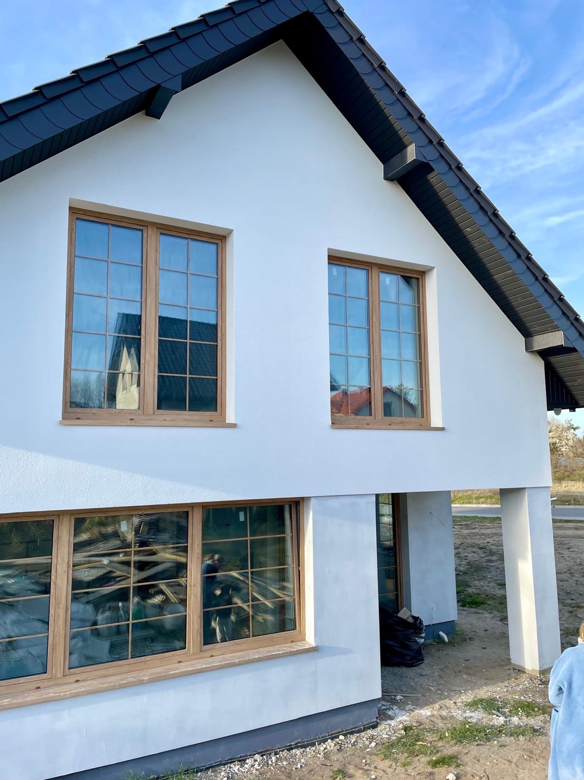 Nowoczesny dom z białymi ścianami i drewnianą stolarką okienną, czarnym dachem. W oknach odbija się otoczenie i wnętrze budynku. Widoczne kolumny podtrzymujące.