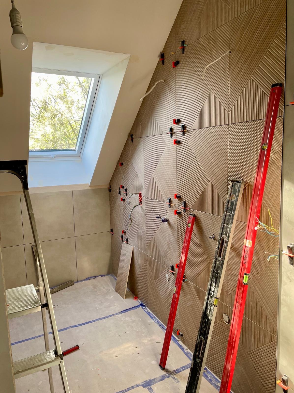 Układanie dekoracyjnych płytek na poddaszu z oknem dachowym. Poziomice i krzyżyki dystansowe w użyciu. Widoczny etap prac wykończeniowych w łazience.