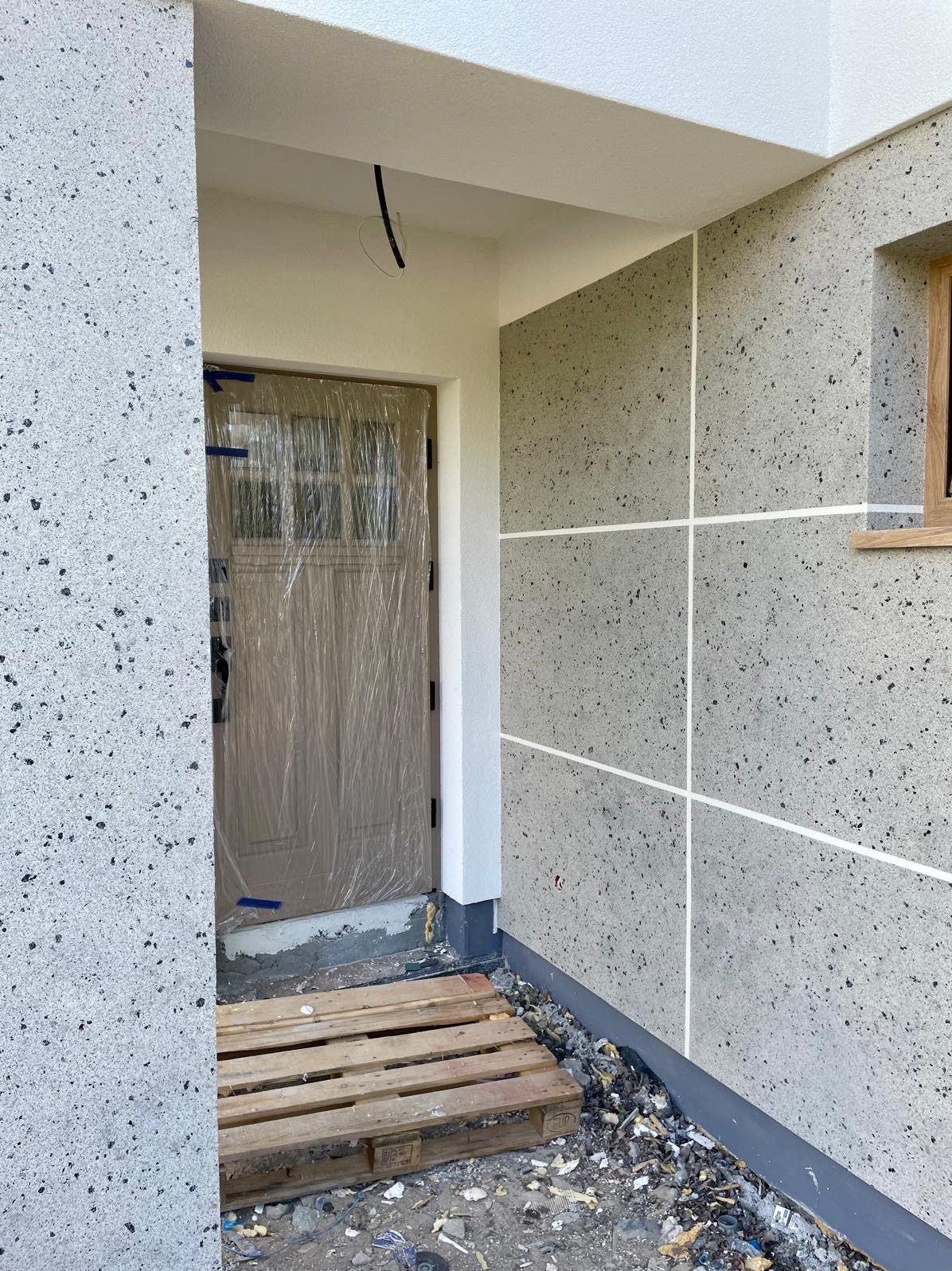 Wejście do domu z elewacją w kropki. Drzwi zabezpieczone folią, widoczna paleta. Fragment okna po prawej. Elewacja podzielona białymi liniami.