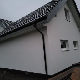 Jakub Czech - Boczny widok domu z białą elewacją i ciemną dachówką. Widoczne okna, rynny i fragment terenu wokół budynku. Pochmurne niebo w tle.