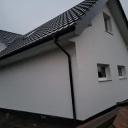 Boczny widok domu z białą elewacją i ciemną dachówką. Widoczne okna, rynny i fragment terenu wokół budynku. Pochmurne niebo w tle.